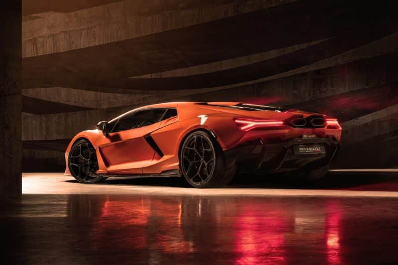Açısal bir tasarıma ve parlayan arka lambalara sahip şık bir turuncu Lamborghini Revuelto, en son özelliklerini sergileyen karanlık, dokulu bir arka plana karşı yansıtıcı bir zemine park edilmiştir.